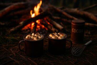 Rustic pot of hot chocolate topped with toasted marshmallows cooking over a campfire.