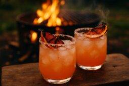 A smoky grapefruit paloma cocktail in a glass with a charred grapefruit garnish by a campfire.