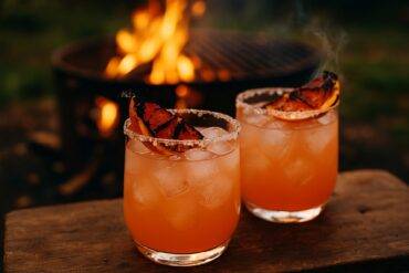 A smoky grapefruit paloma cocktail in a glass with a charred grapefruit garnish by a campfire.